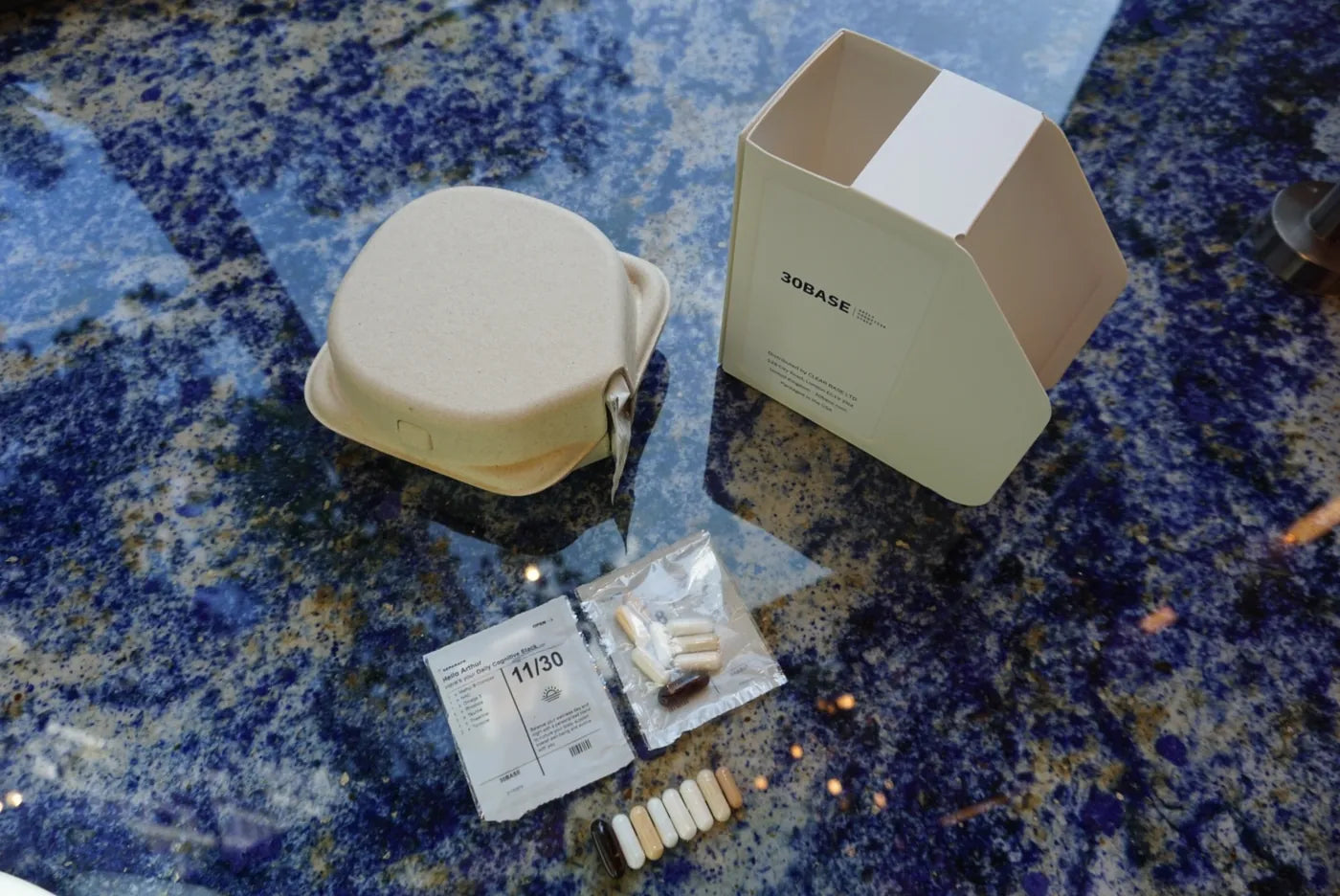 An open 30BASE box on a marble ledge by a garden, the dated daily sachets visible inside.