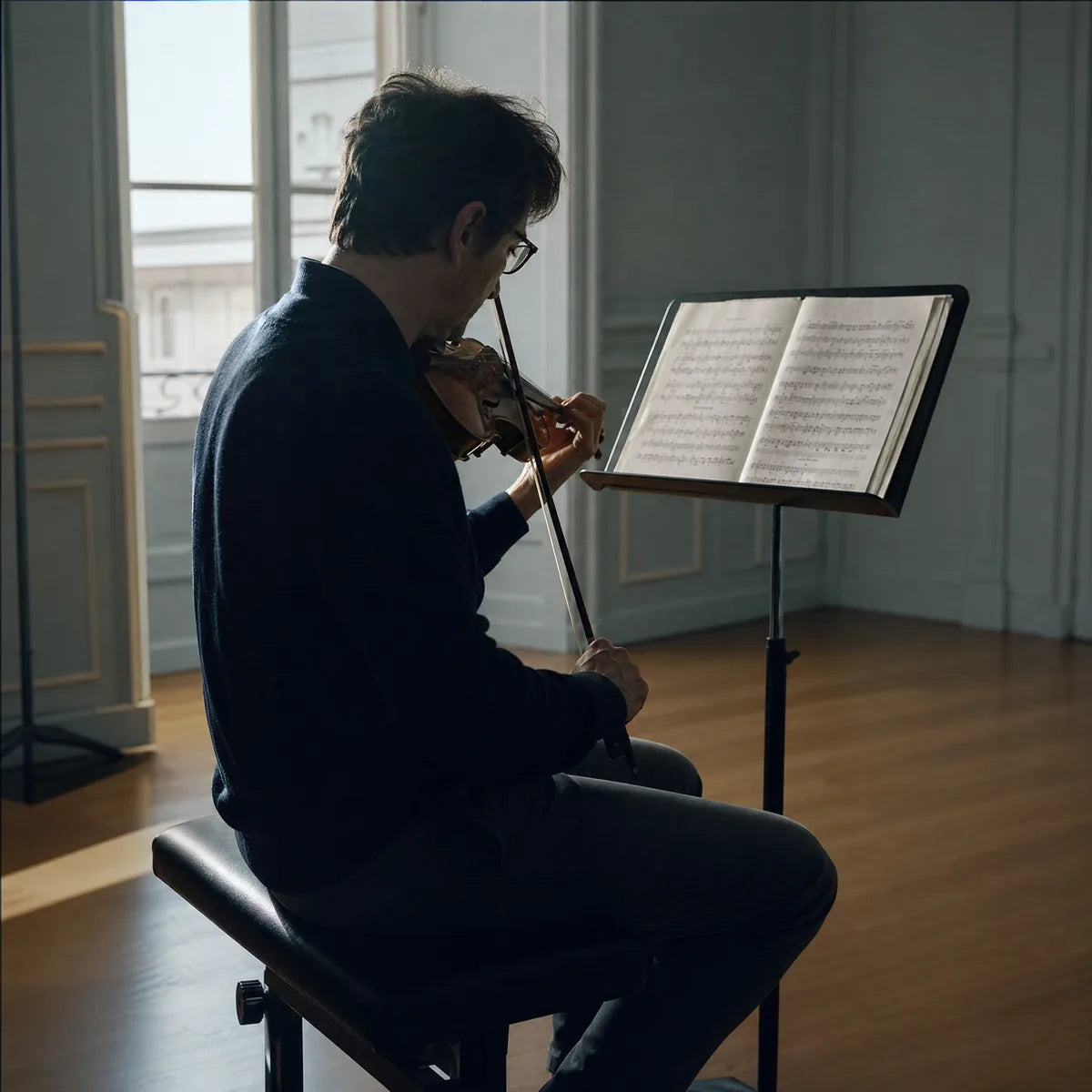 A musician reading a score and playing the violin beside a tall window.