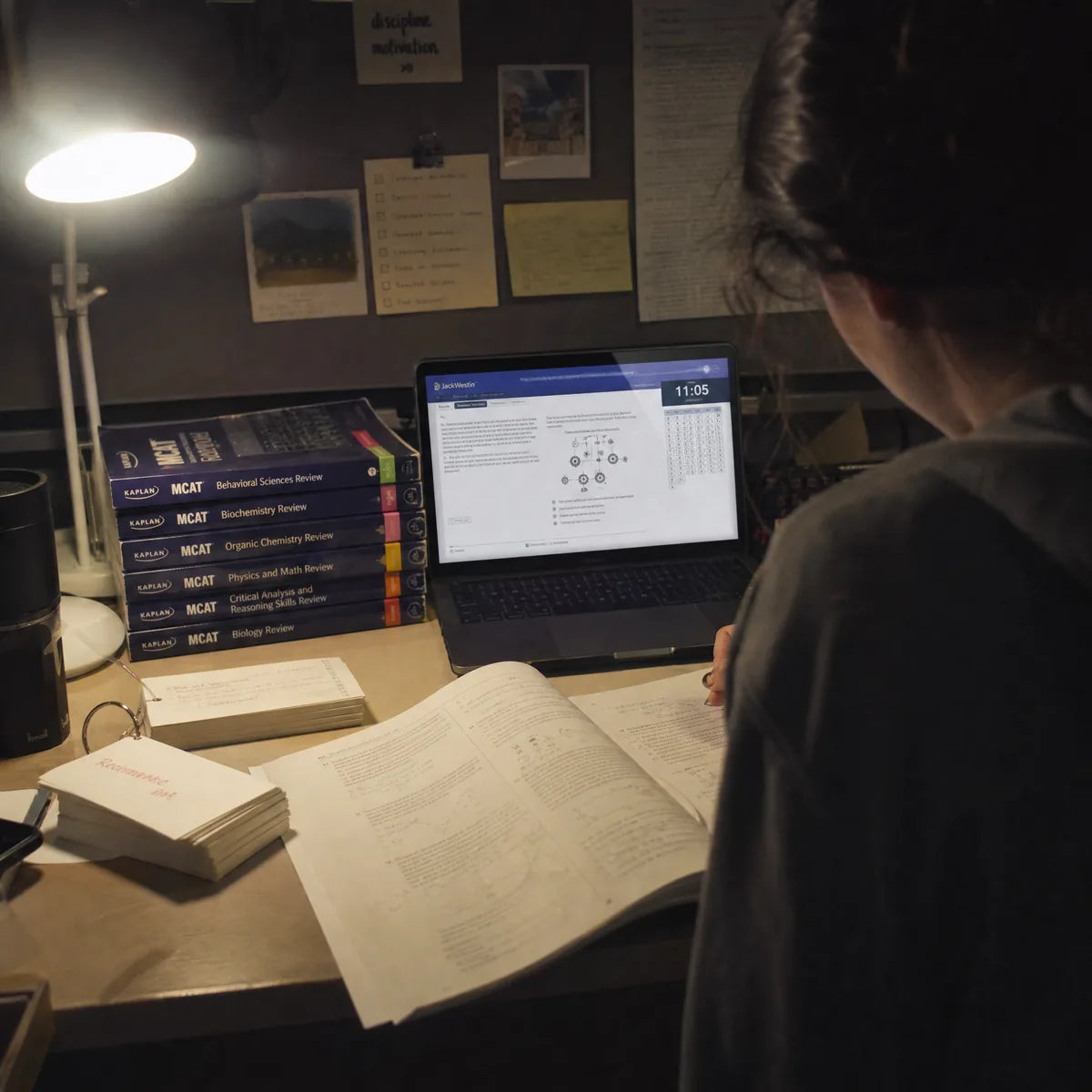 A student preparing for an exam at a desk with textbooks, flashcards and a laptop under a warm lamp.